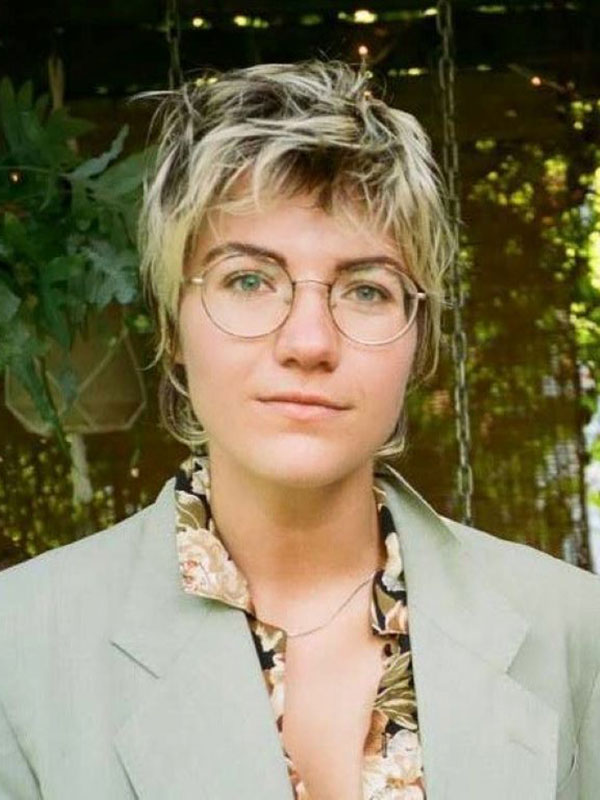 A headshot of Maggie McLaughlin, who wears a beige blazer and poses near hanging plants and an enclosure canopied with vegetation.