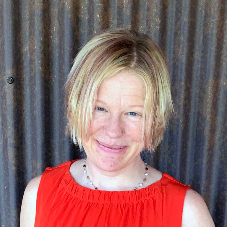 A headshot of Professor Jennifer Goodlander, who wears a red shirt and poses against a corrugated metal background.