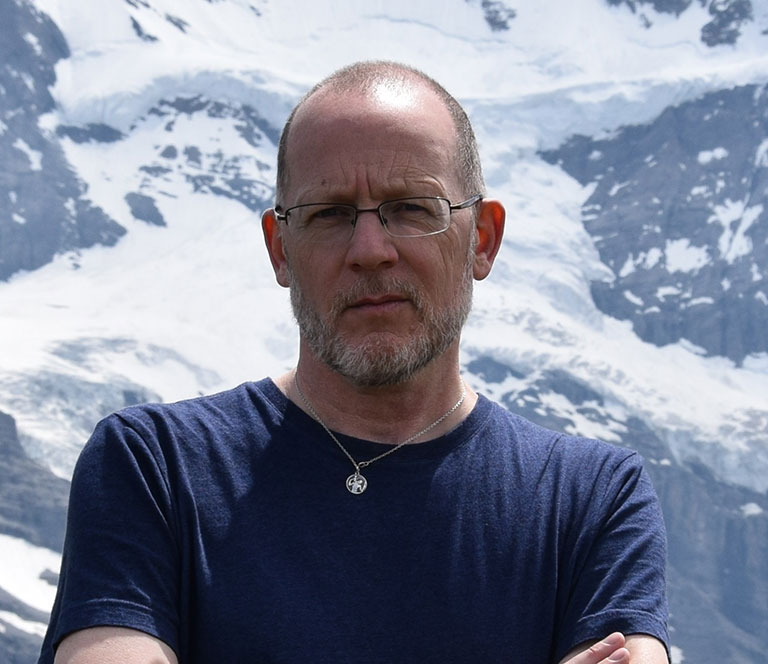 A photo of Professor Bill Johnston, who poses outside with a snowy mountain in the background.
